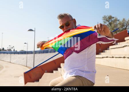Handsome mature man with beard and grey hair is sitting on the bleachers of an open-air auditorium while holding the gay pride flag in his hands. Conc Stock Photo