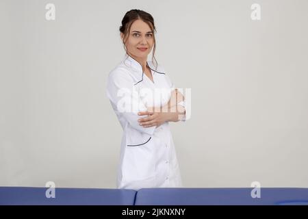 Smiling, glad woman doctor in medical uniform standing near blue massage table and looking at camera with crossed arms Stock Photo
