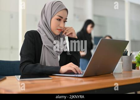 Professional millennial Asian Muslim businesswoman or female manager concentrating on her work project on laptop computer. Stock Photo