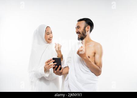 Excited couple wearing ihram clothes using cellphones with clenched hands Stock Photo