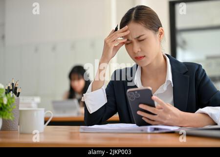 Unhappy young Asian businesswoman or female office worker at her office desk suffering from a headache, being overworked, and having a career problem. Stock Photo