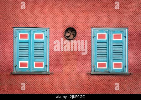 Two painted closed window shutters and a fan on an orange colored wall of a building Stock Photo