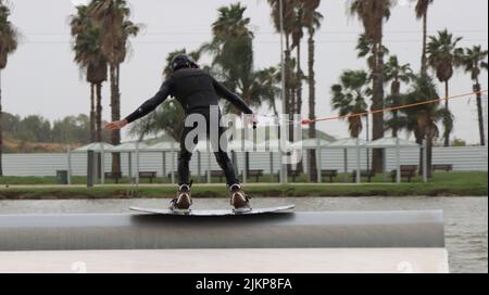 A back shot of a male on a skateboard holding a rope connected to a boat with palm trees in the background Stock Photo