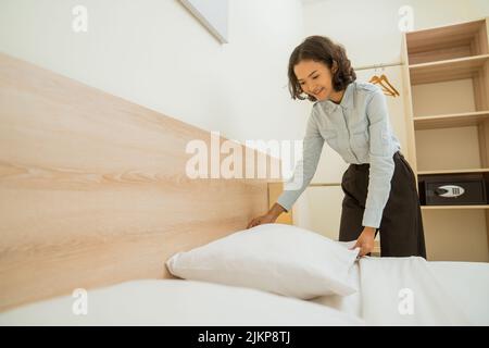 young female housekeeper tidying pillows on bed in hotel room Stock Photo