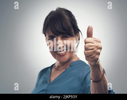 Well done on your sterling work. Studio portrait of a senior woman showing thumbs up against a grey background. Stock Photo