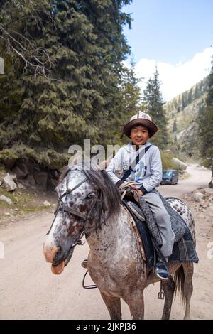 27.07.2022, Kyrgyztan, Issyk-Kul. A cute, smiling Asian boy in a national hat riding a horse rides in the mountains and meets tourists. Close-up. Stock Photo