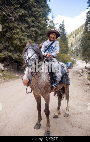 27.07.2022, Kyrgyztan, Issyk-Kul. A cute, smiling Asian boy in a national hat riding a horse rides in the mountains and meets tourists. Close-up. Stock Photo