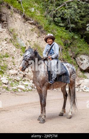 27.07.2022, Kyrgyztan, Issyk-Kul. A cute, smiling Asian boy in a national hat riding a horse rides in the mountains and meets tourists. Close-up. Stock Photo