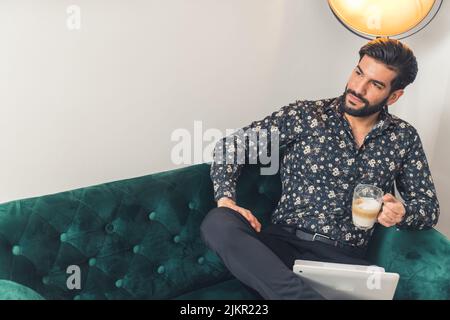 Young hispanic man wearing shirt sitting in living room working on laptop taking a break to drink coffee looking to the side. Indoor shot. High quality photo Stock Photo