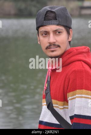 Waist-up of a Indian young guy wearing cap backwards looking sideways while standing by the lake Stock Photo