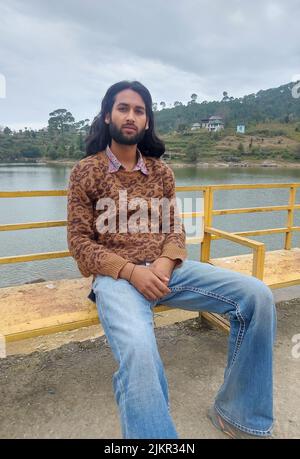A good looking Indian young man with long hair and beard looking at camera while sitting on bench beside the lake Stock Photo