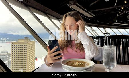 a teenage girl in a white shirt with blond hair sits and looks at the iPhone 13 phone she is interested And with her finger something corrects the black phone Top Of Vancouver Revolving Restaurant. Stock Photo
