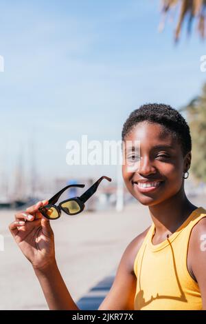 portrait of a positive african woman looking at camera near coast. She is wearing summer yellow t-shirt and holds yellow sunglasses Stock Photo