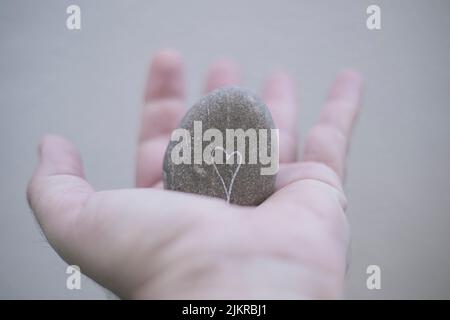 Man's hand holding a gray rounded stone on which there is a white heart drawn in a well-lit environment Stock Photo