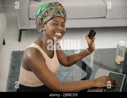 African woman wearing traditional headscarf, making online shopping purchase using laptop and credit card. Sitting by coffee table in lounge at home Stock Photo