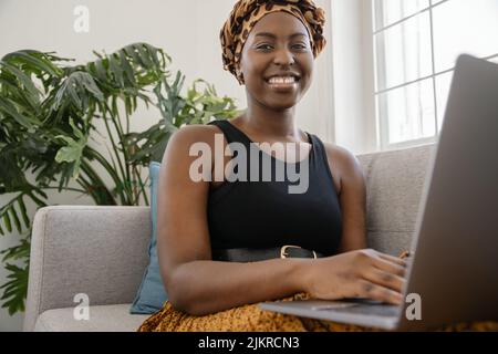 Close up Beautiful traditional young gen z African woman sitting on sofa at home, typing on laptop, smiling, looking at camera Stock Photo
