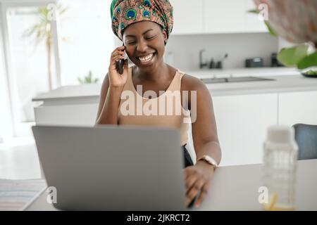 Beautiful young African woman wearing tradition headscarf. Sitting at home working on laptop. On phone call smiling and gesturing with hand Stock Photo