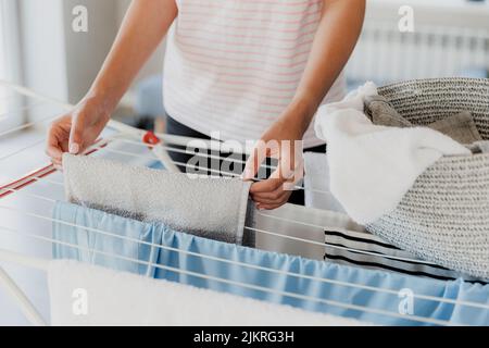 Woman hanging clean wet clothes laundry on drying rack at home laundry room. Detail of female housewife hands closeup holding, spreading and taking laundry basket in front of clothing rack dryer Stock Photo
