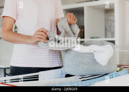 Woman boring, hanging clean wet clothes laundry on drying rack at home. Detail of female housewife hands closeup holding, spreading and taking laundry basket in front of clothing rack dryer Stock Photo