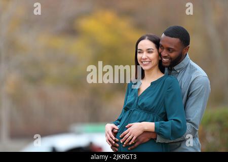Interracial couple looking away enjoying pregnancy in a park Stock Photo