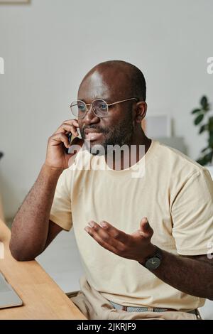 African young businessman in eyeglasses having conversation on mobile phone sitting at table at office Stock Photo