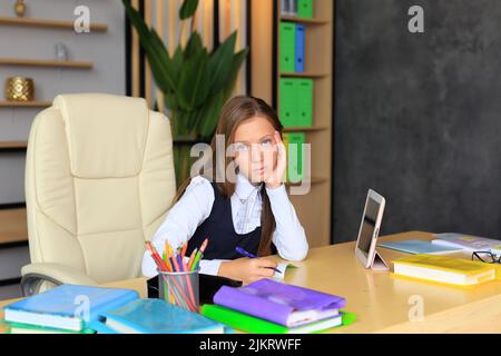 A girl in a school uniform sits at the table for lessons. The child does homework with books and a tablet. Classroom Stock Photo