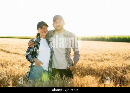 A couple of farmers in plaid shirts and caps stand embracing on agricultural field of wheat at sunset  Stock Photo