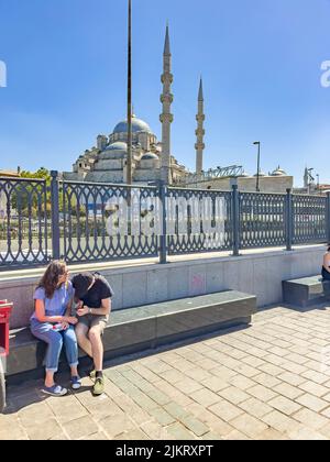 Tourist couple sitting at a bench in front of Yeni Cami, New Mosque, at Eminonu, Sirkeci in Istanbul old downtown district and checking their mobile Stock Photo