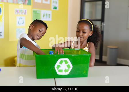 African american elementary students putting bottles in recycling container at desk in school Stock Photo