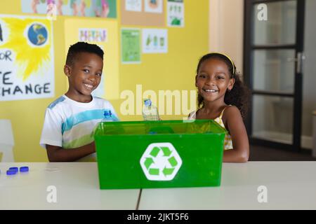 Portrait of smiling african american elementary students with recycling container at desk in school Stock Photo