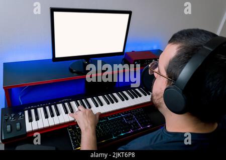 side top view young caucasian man with beard, headphones and glasses making musical arrangements with a synthesizer piano, recording in his home studi Stock Photo