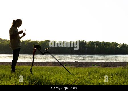 Silhouette of a guy who puts on wireless headphones.  In front of him stands a wireless metal detector supported by a shovel.  On the river bank, fore Stock Photo