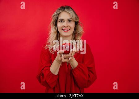 Young cheerful, merry, blissful blonde woman hold and present to camera fresh organic apple in two hands in red studio Stock Photo