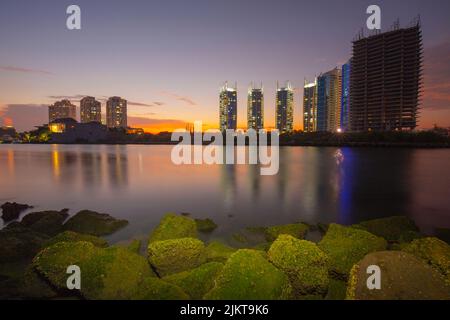 A scenic view of mossy rocks and calm sea against city buildings during sunset Stock Photo