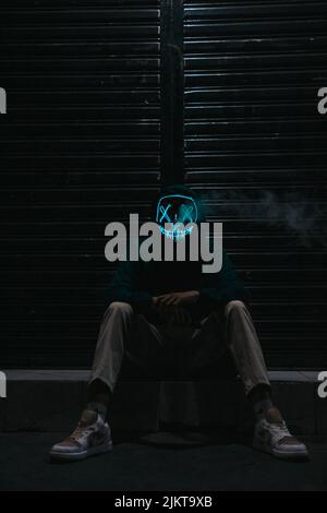 A vertical shot of man sitting in street wearing a neon mask Stock Photo