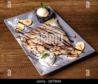 A plate of chocolate crepe with chocolate sauce drizzles, whipped cream banana slices on a wooden table Stock Photo