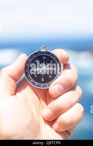 Closeup, hand holding an analog compass in front of the sea, summer concept, holidays, travelling. Selective focus, vertical shot. Stock Photo