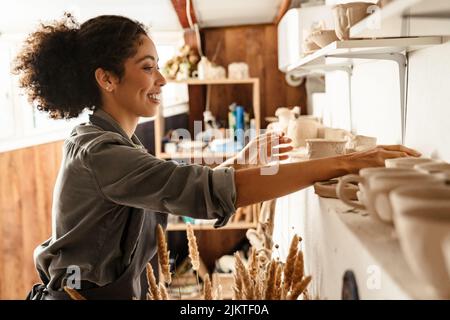 Young black ceramist woman wearing apron smiling while working in her studio Stock Photo