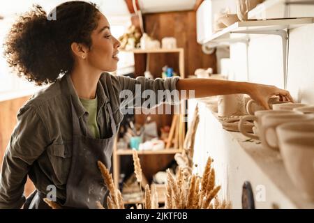 Young black ceramist woman wearing apron smiling while working in her studio Stock Photo