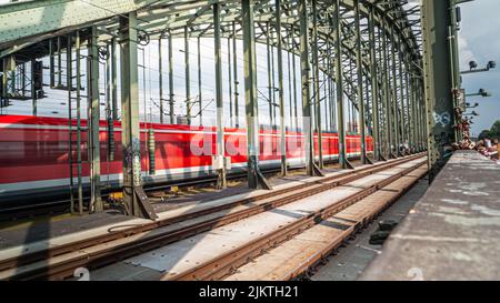 A view of a railway station with a red train moving and the surrounding structures on a sunny day Stock Photo