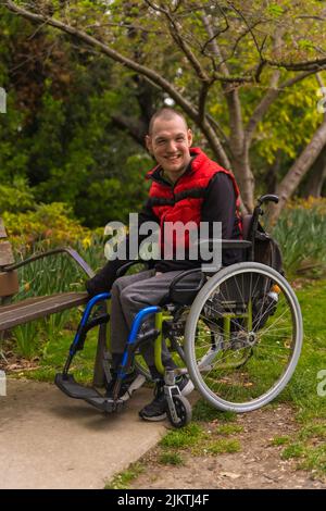 Portrait of a paralyzed young man in a red vest in a public park in the city. Sitting in the wheelchair next to a park chair Stock Photo