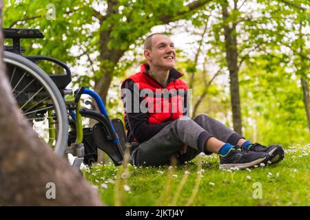 Portrait of a paralyzed young man sitting on the grass next to the wheelchair next to a tree Stock Photo