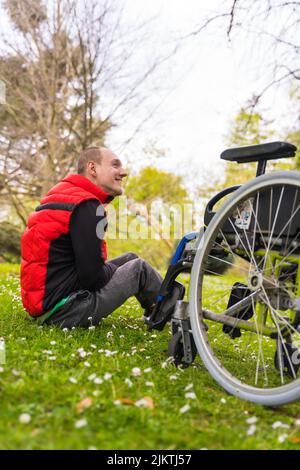 Paralyzed young man sitting on the grass next to the wheelchair next to a tree looking at the camera and smiling Stock Photo