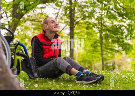 Portrait of a paralyzed young man sitting on the grass next to the wheelchair next to a tree Stock Photo
