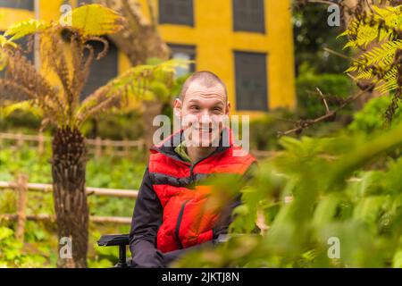Portrait of a paralyzed young man in a public park in the city. Sitting in the wheelchair smiling Stock Photo