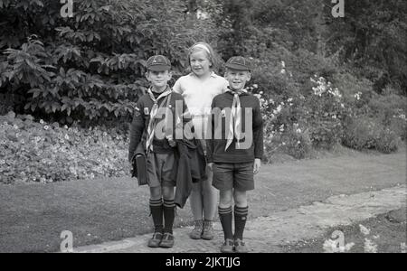 1960s, historical, outside on a garden path, a girl standing for a photo with her two younger brothers, both wearing their Wolf cub scout uniform of the era, England, UK. Known as Wolf cubs a name originally derived from the Kipling story, The Jungle Book later in this era, young boy scouts began to become known simply as Cubs or Cub scouts, while retaining the traditional Wolf cub ceremonies such as the Grand Howl. Stock Photo