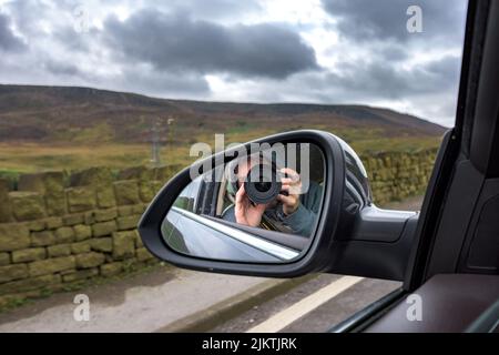 A man taking a photo with a camera in the side mirror of a car and a cloudy sky in the background Stock Photo