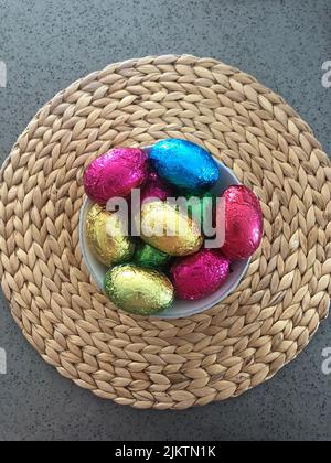 from above colourful foil wrapped chocolate easter eggs in a bowl with straw wicker woven placemat on a polished concrete bench top Stock Photo