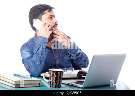young man sitting in front of a laptop thinking and talking on cell phone in white background Stock Photo