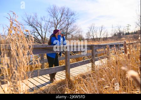 A woman in a blue down jacket looks into the distance through blue binoculars on a bridge in a park on a sunny spring day. Stock Photo